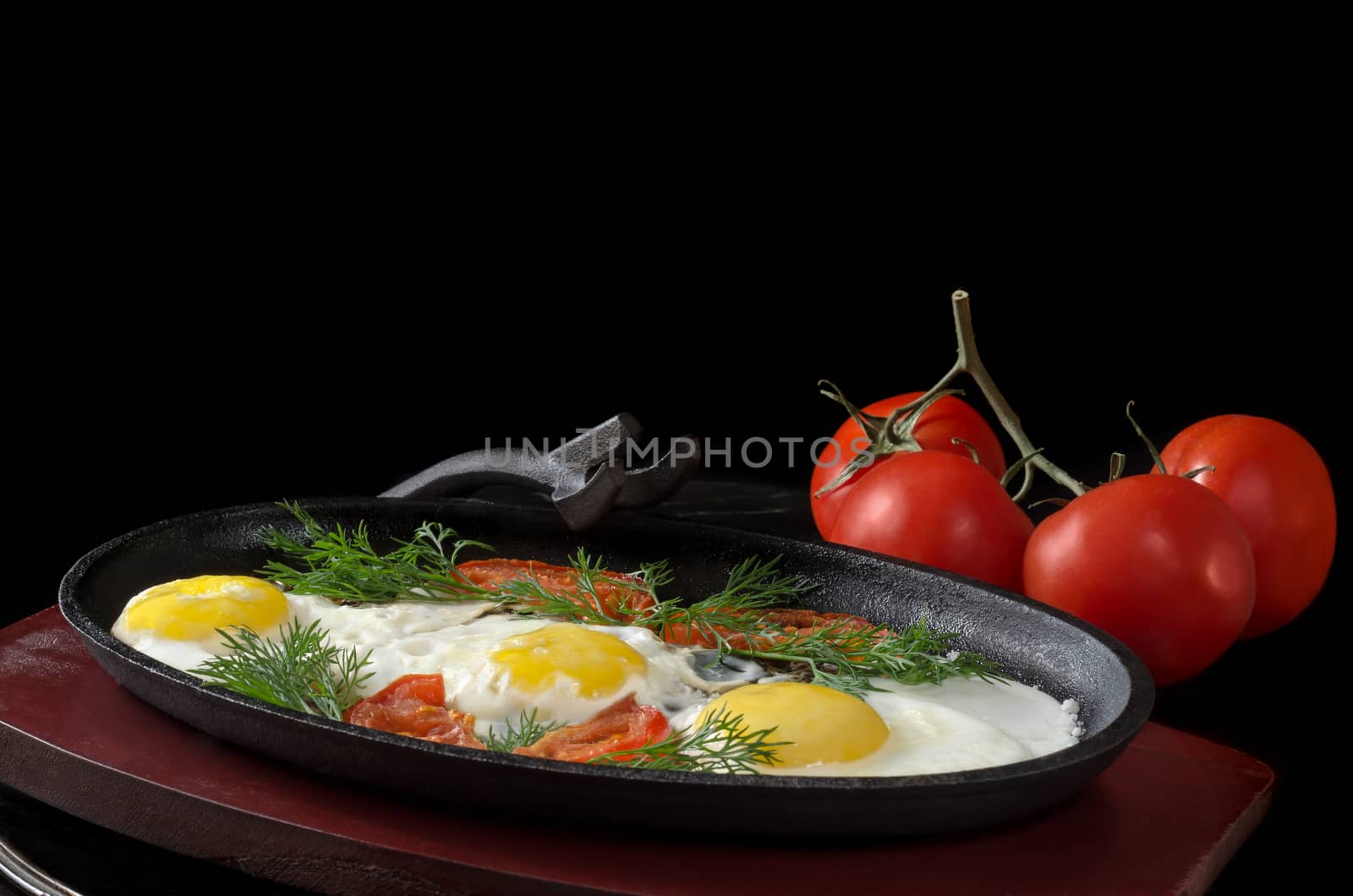 Scrambled eggs with tomatoes in a frying pan on the stand,and the whole tomatoes on black background
