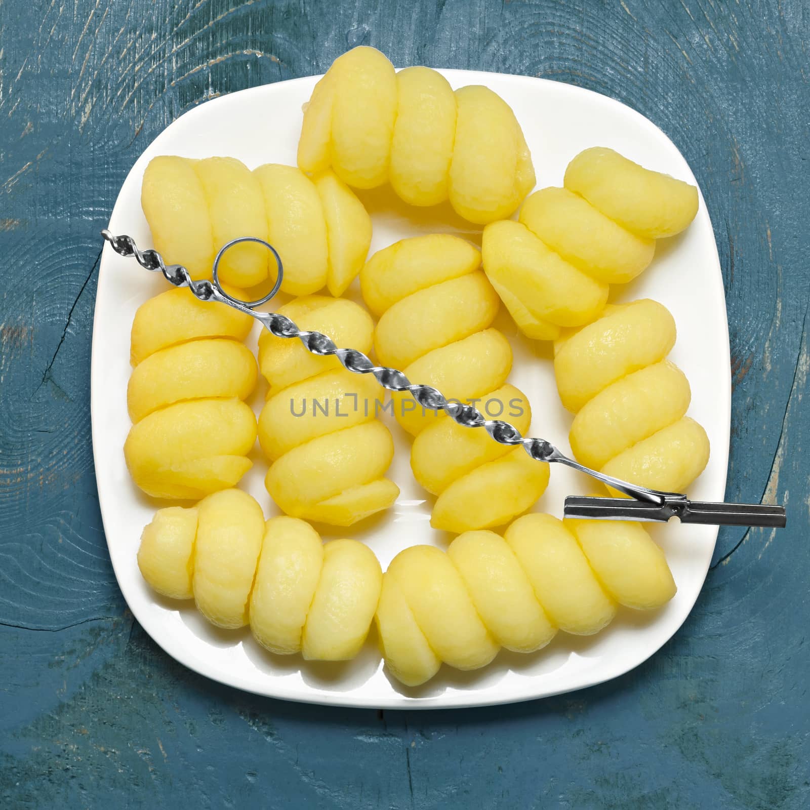 Raw potatoes, sliced, shaped, lies on a plate on blue wooden background, and tool for cutting