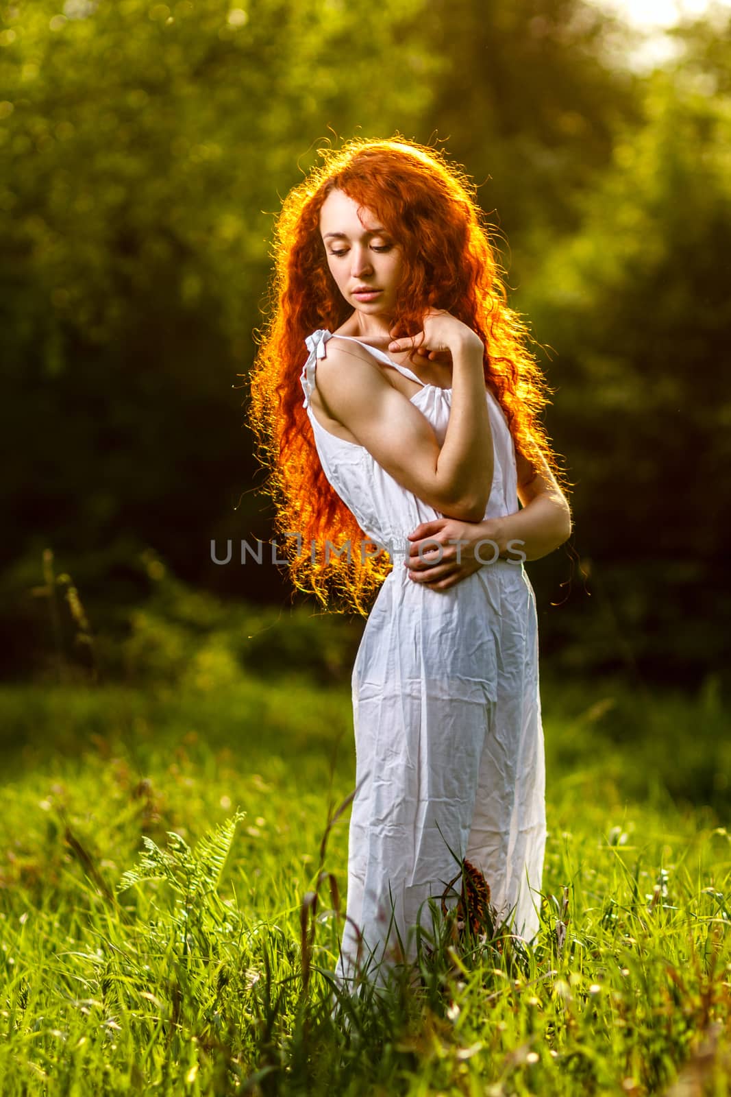 Beautiful redhead girl with curly hair walking under the rays of the sunset in summer spring forest wearing dress