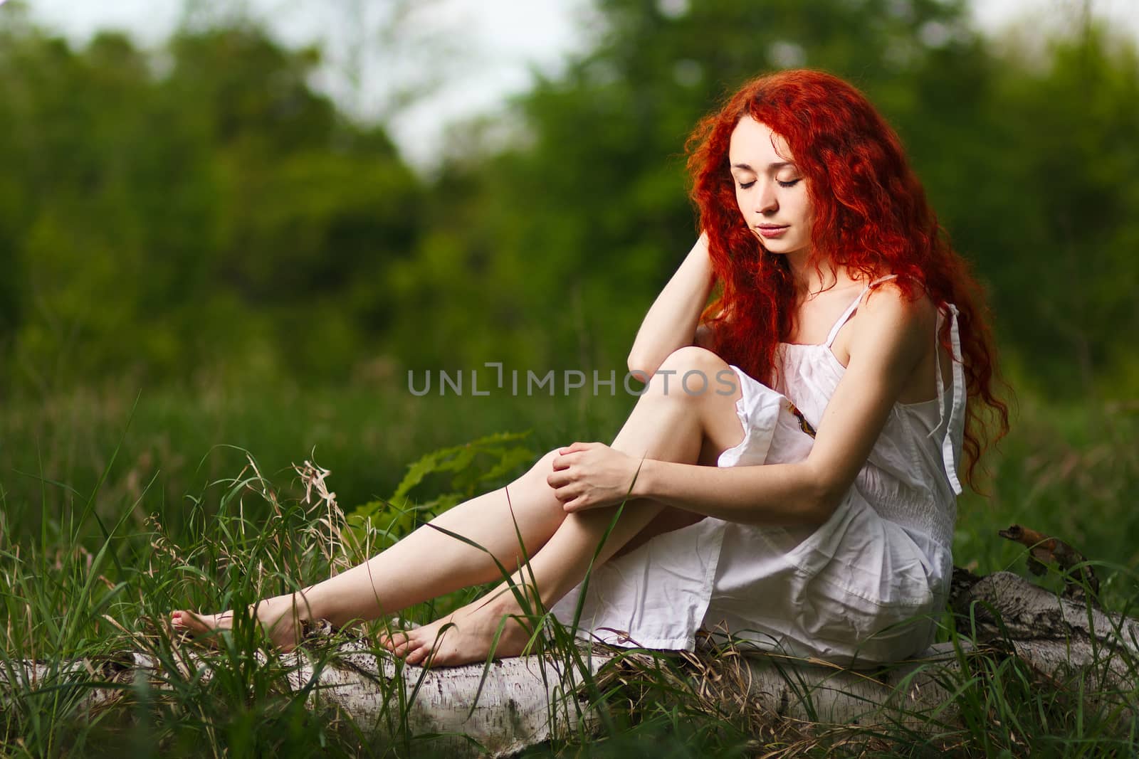 Beautiful redhead girl with curly hair walking under the rays of the sunset in summer spring forest wearing dress