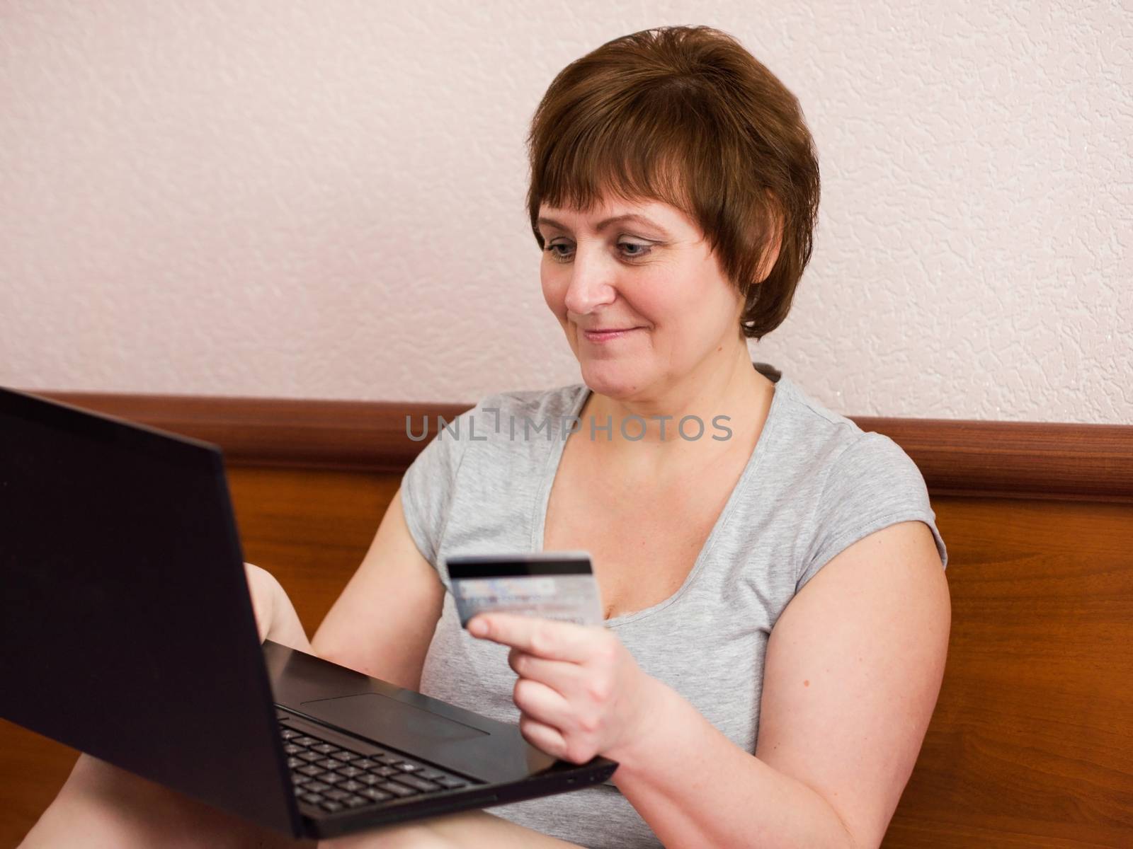 Smiling senior adult woman sitting at bedroom with laptop and holding bank card in her hand
