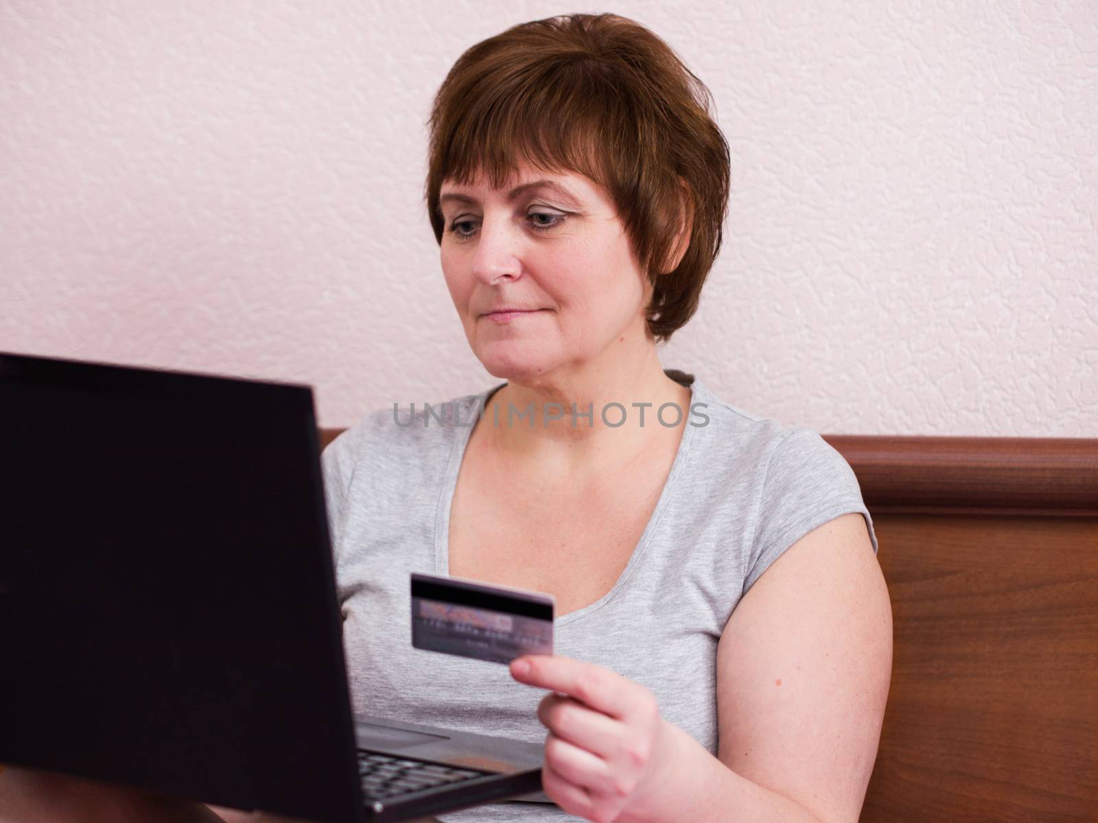Portrait of senior woman sitting at bedroom with her laptop and  holding bank card in her hand, senior adult woman makes a purchase online.