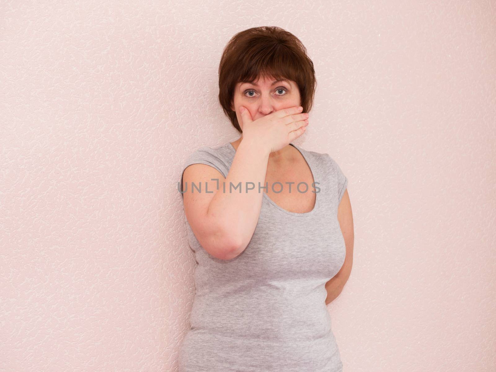 Portrait attractive mature woman with shocked, surprised, anxious facial expression, covering mouth with hand on plain background