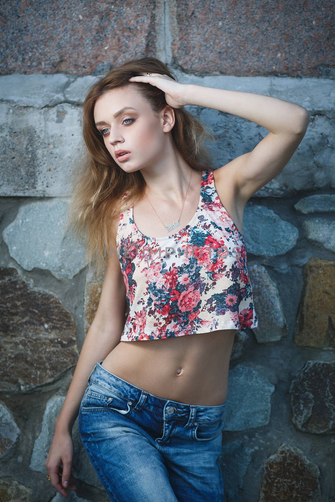 Young sexy woman model in jeans and tank top posing for fashion portrait near old weathered wall outside