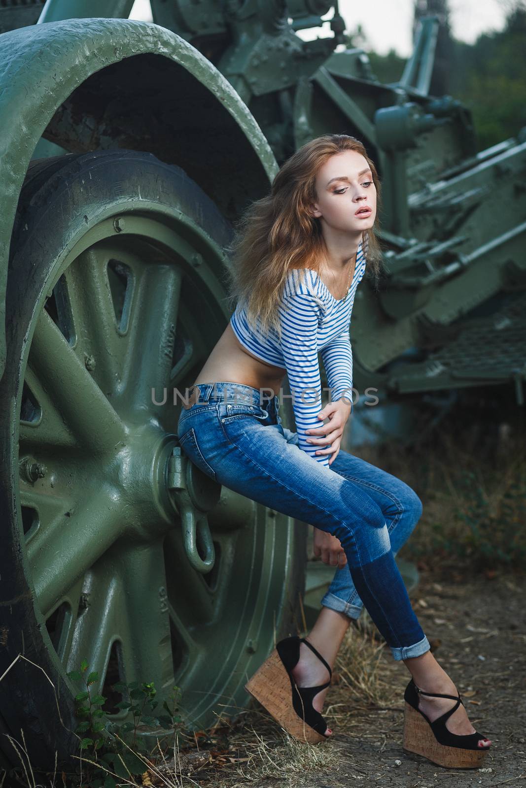 Young sexy woman model in jeans and striped tank top posing for fashion portrait near old artillery gun outside