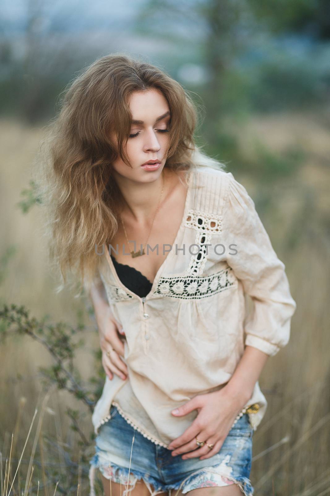 Young sexy woman model in jeans shorts and blouse posing for fashion portrait outside in a field at golden hour sunset