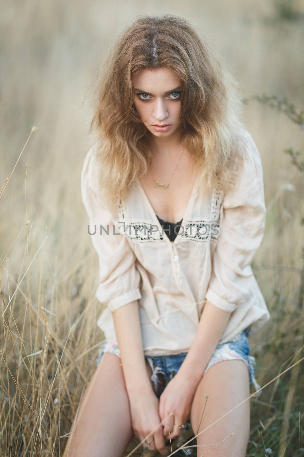 Young sexy woman model in jeans shorts and blouse posing for fashion portrait outside in a field at golden hour sunset