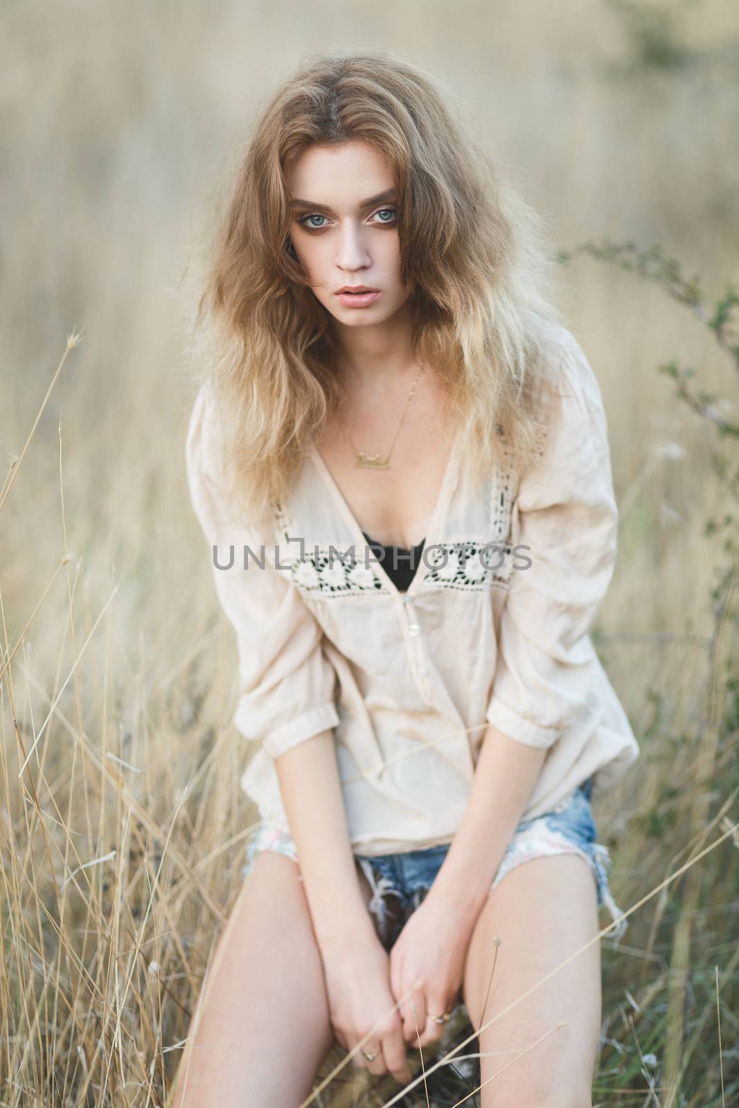 Young sexy woman model in jeans shorts and blouse posing for fashion portrait outside in a field at golden hour sunset