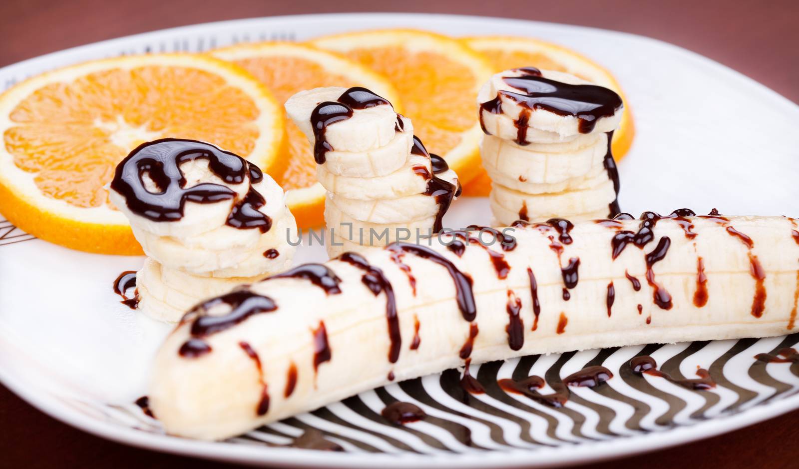 Fresh fruit dessert with banana and orange topped with melted chocolate on white plate