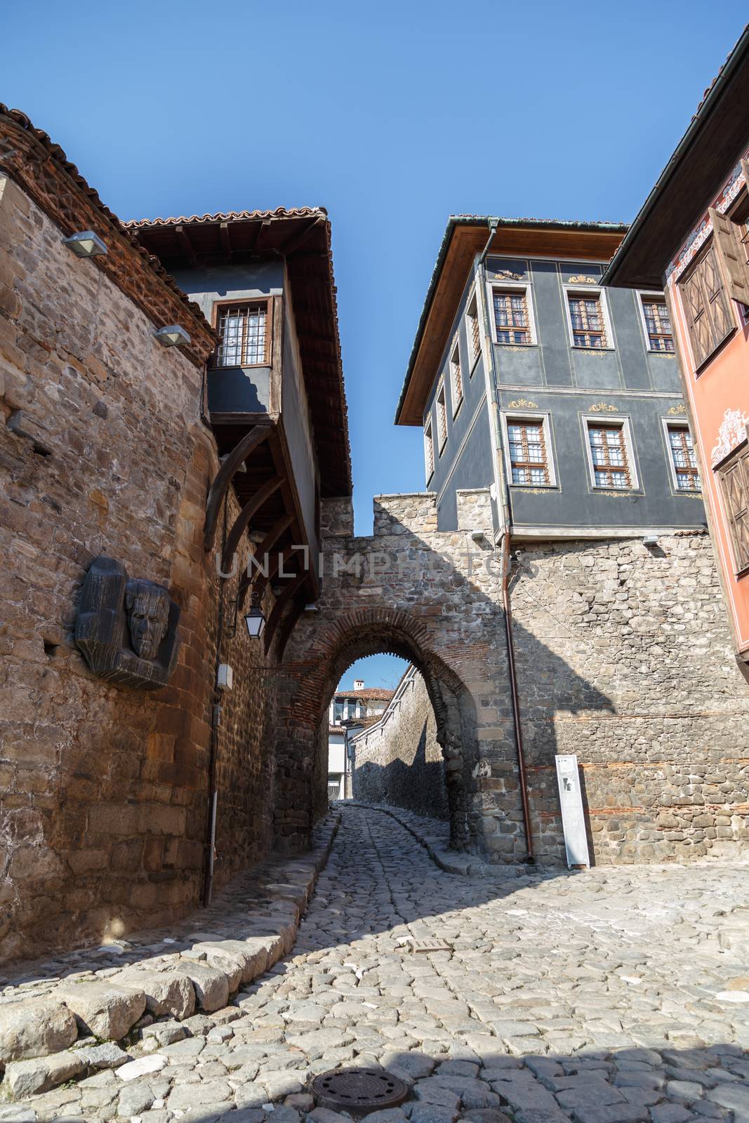 Front view of historical Hisar Gate with pathway and brown and blue old buildings around in Plovdiv old town, on bright blue sky background.