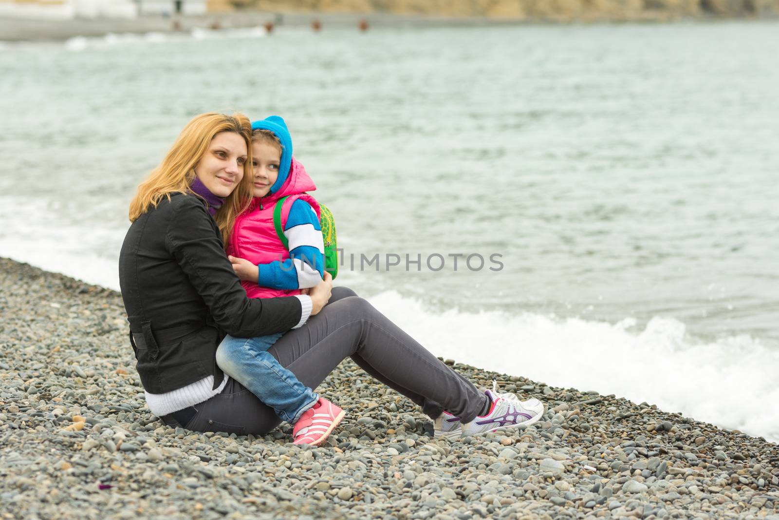 Mom and daughter in the arms sit on the beach on a cool day and watching the surf