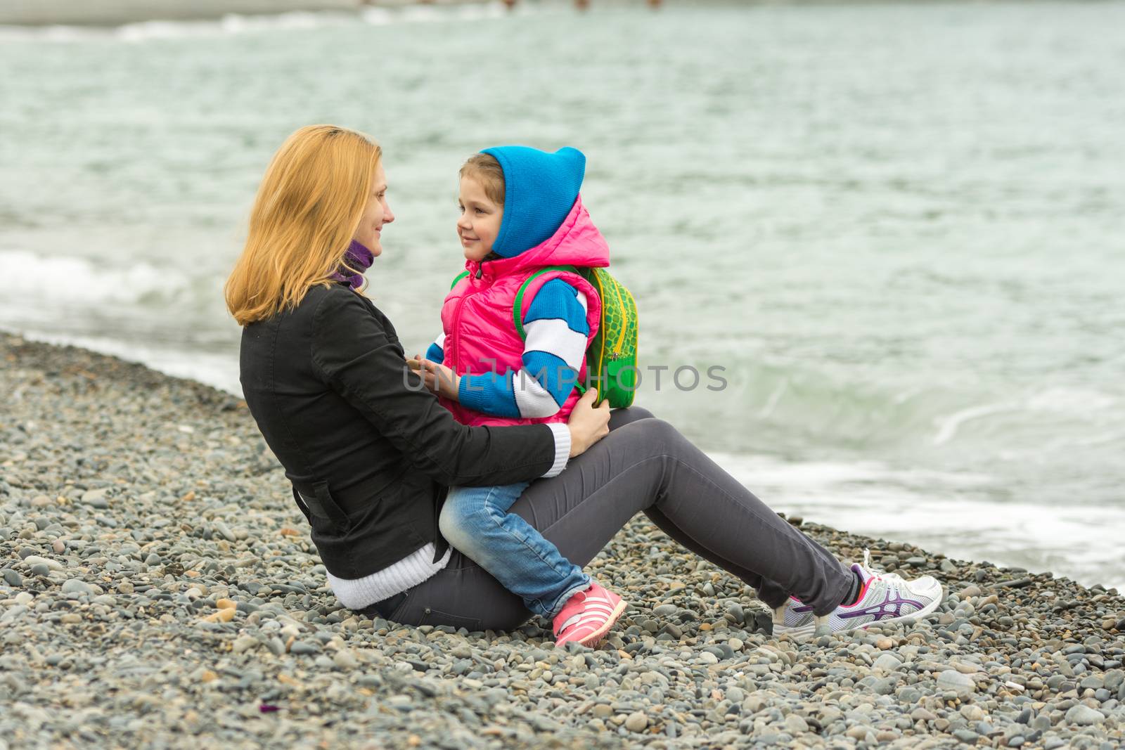 Mother and daughter tenderly looking at each other sitting on the beach on a cool day