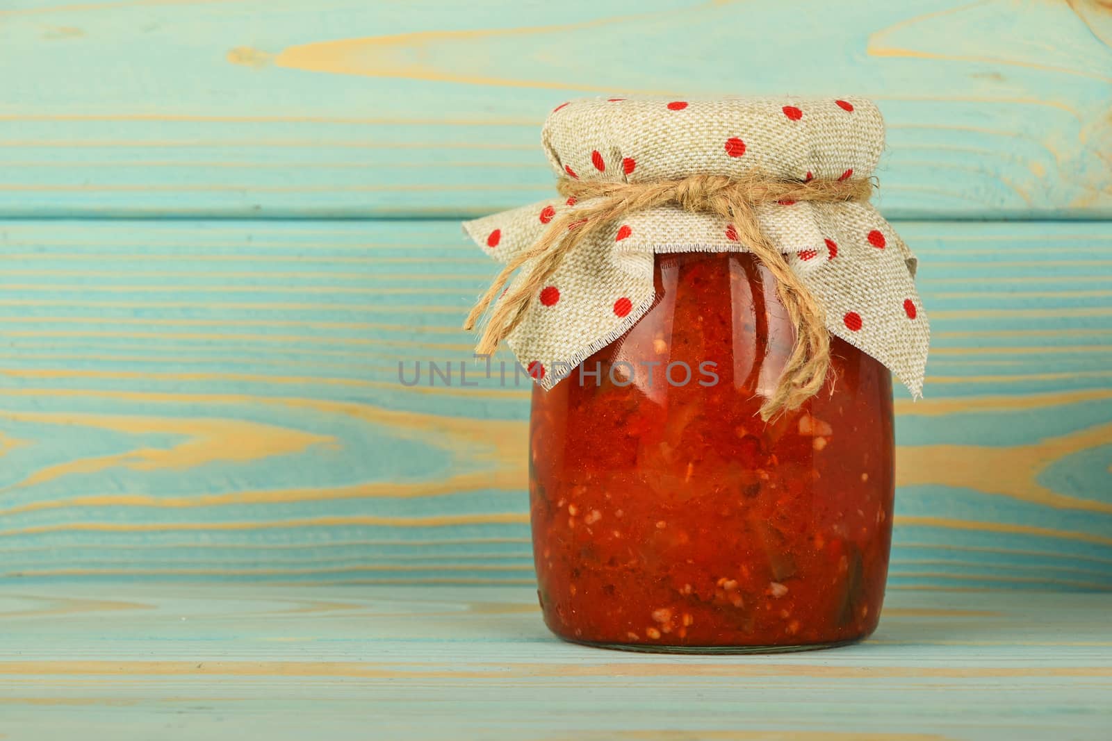 One glass jar of homemade pickled pepper, paprika and eggplant salad with dotted textile top decoration at blue painted vintage wooden surface