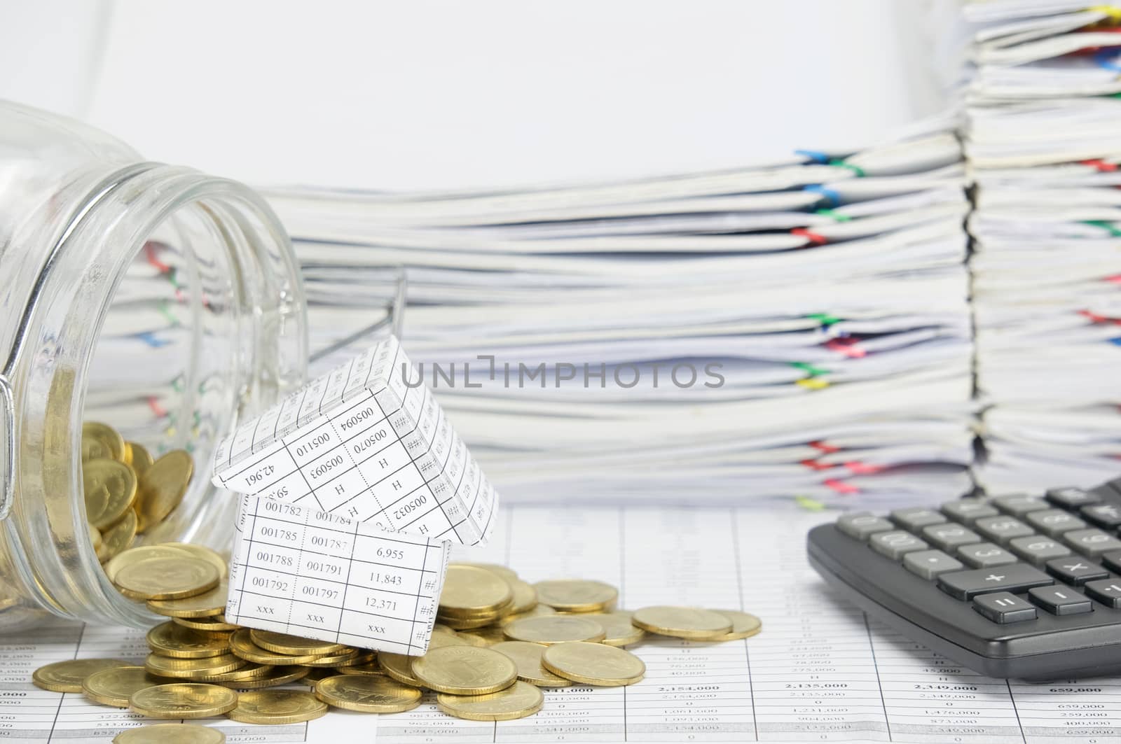 House with stack of gold coins flowing out of bottle on finance account have calculator and overload of paperwork as background.