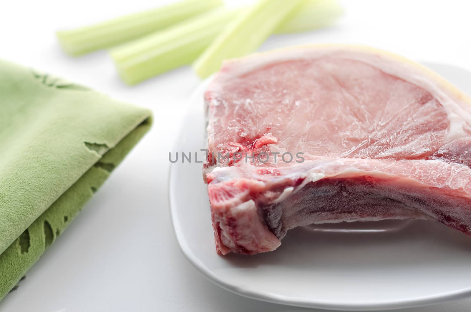 Fresh pork meat on white plate, morning light and bokeh. Green napkin and celery, white background.