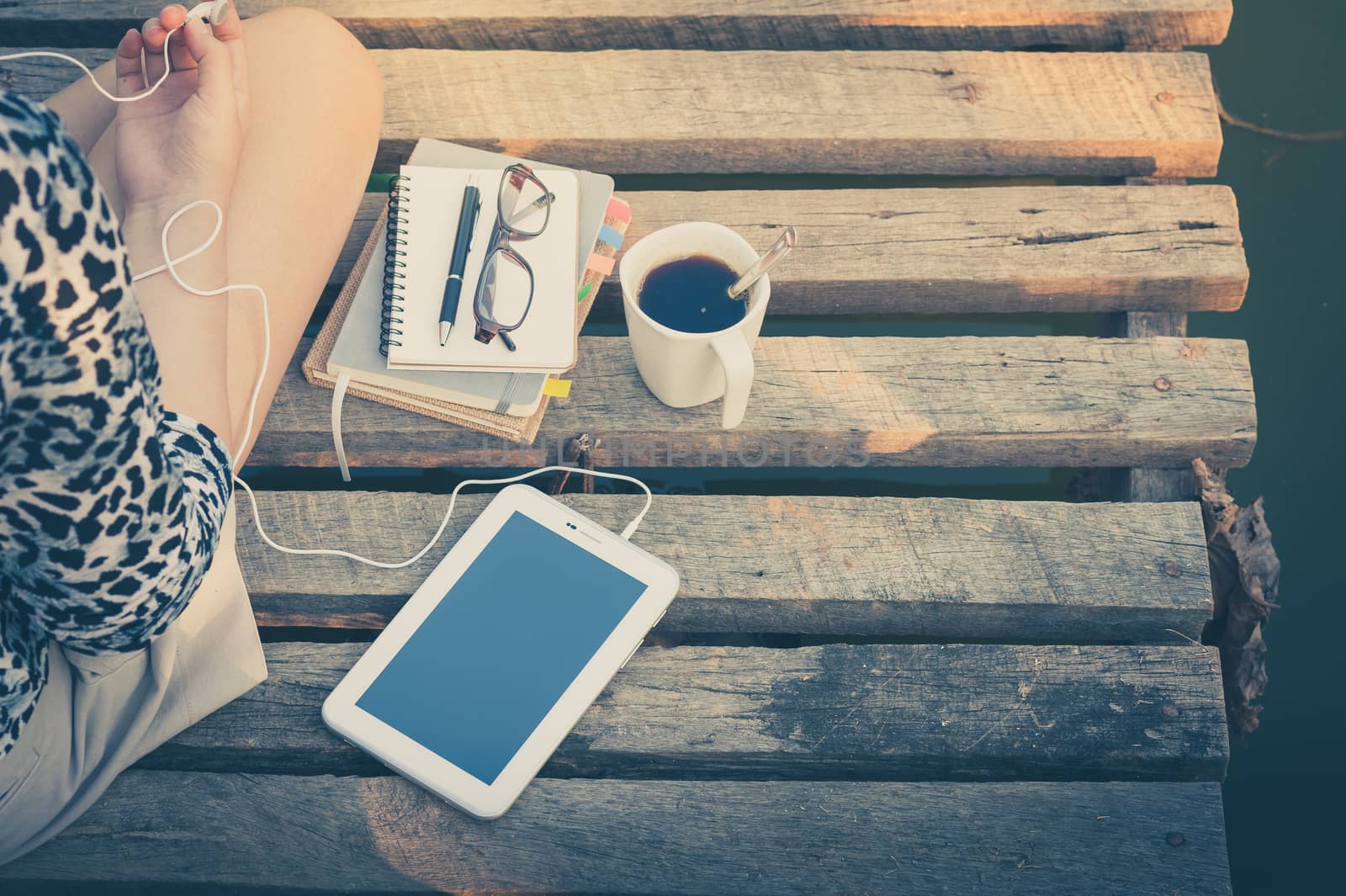 Young woman sitting on wood bridge with small tablet pc, coffee cup, glasses, and notebooks while listening to music in morning time with vintage filter effect