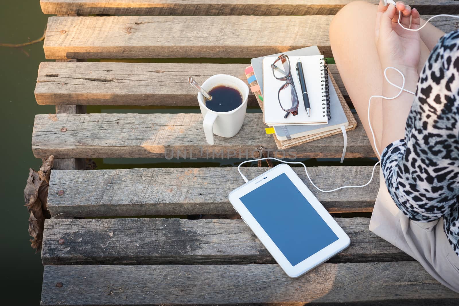 Young woman sitting on wood bridge with small tablet pc, coffee cup, glasses, and notebooks while listening to music in morning time