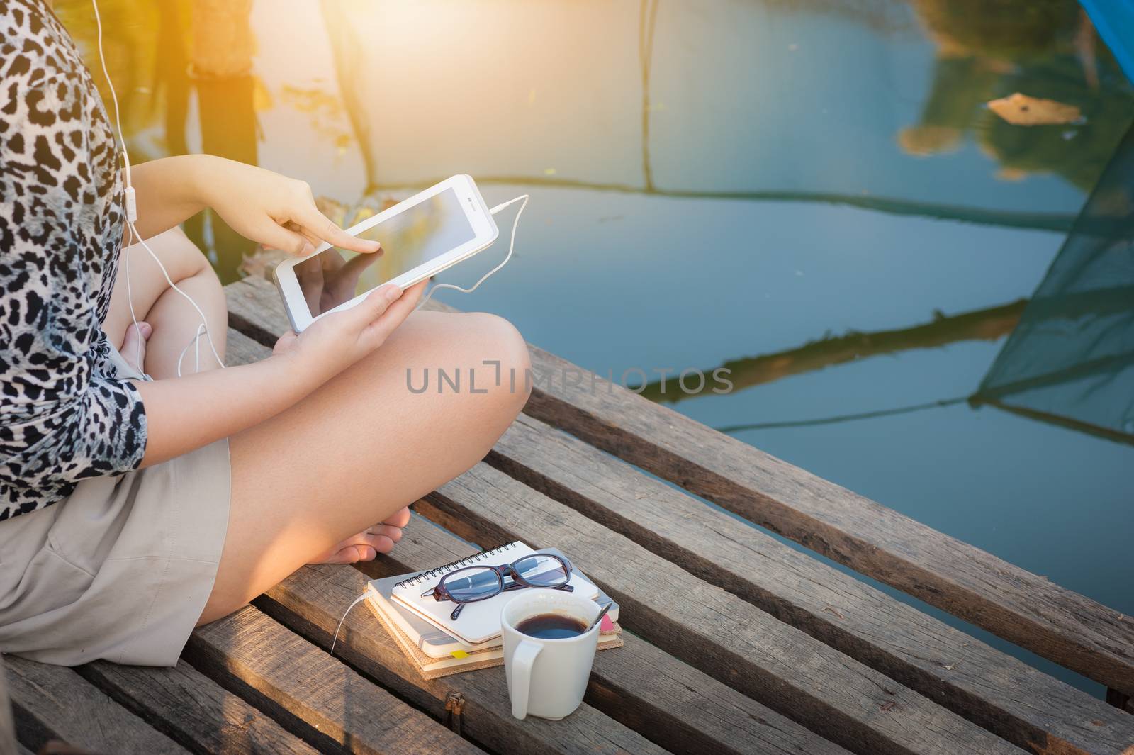 Young woman sitting on wood bridge touching on small talet pc screen while listening to music in morning time on weekend