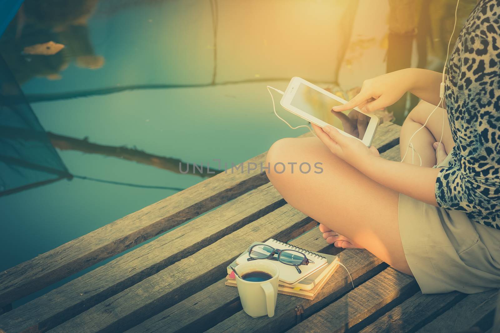 Young woman sitting on wood bridge touching on small talet pc screen while listening to music in morning time on weekend with vintage filter effect