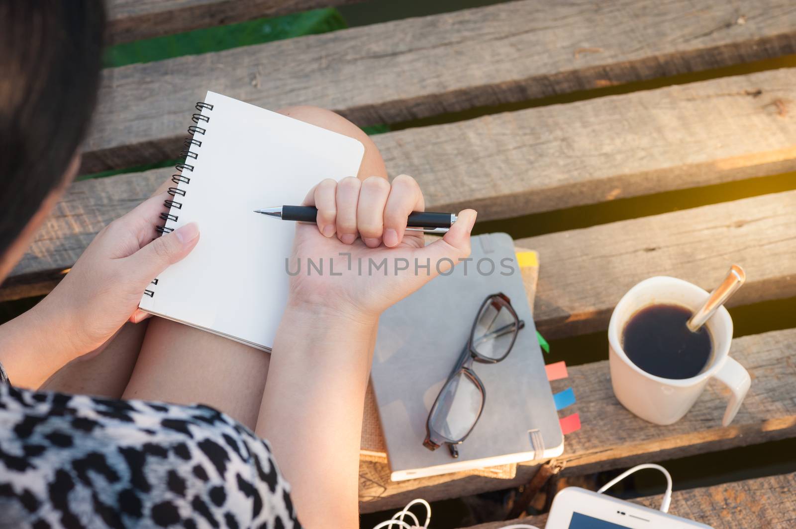 Young hipster woman writing journal on small notebook while sitting on wood bridge in morning time on weekend