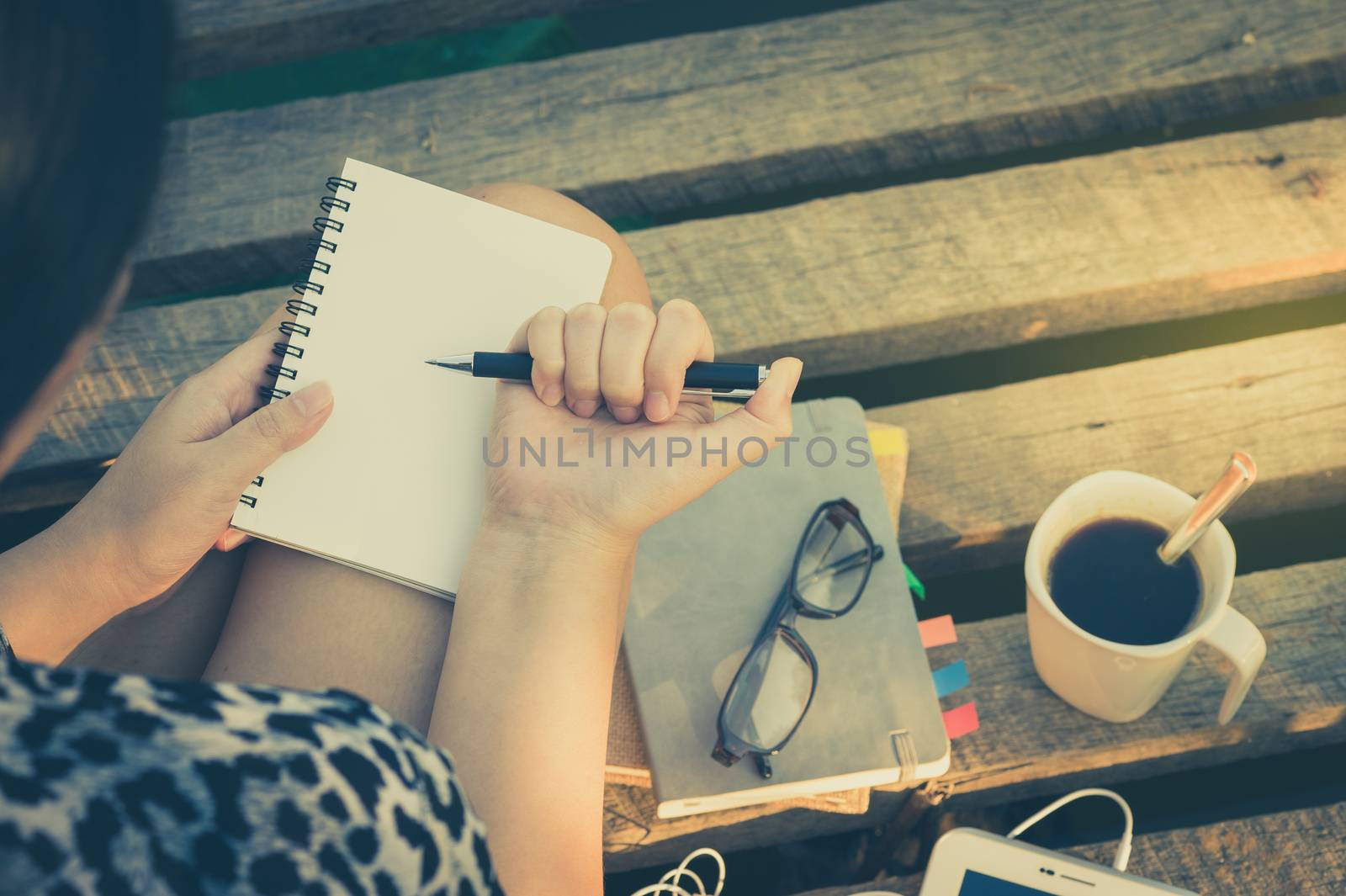 Young hipster woman writing journal on small notebook while sitting on wood bridge in morning time on weekend with vintage filter effect