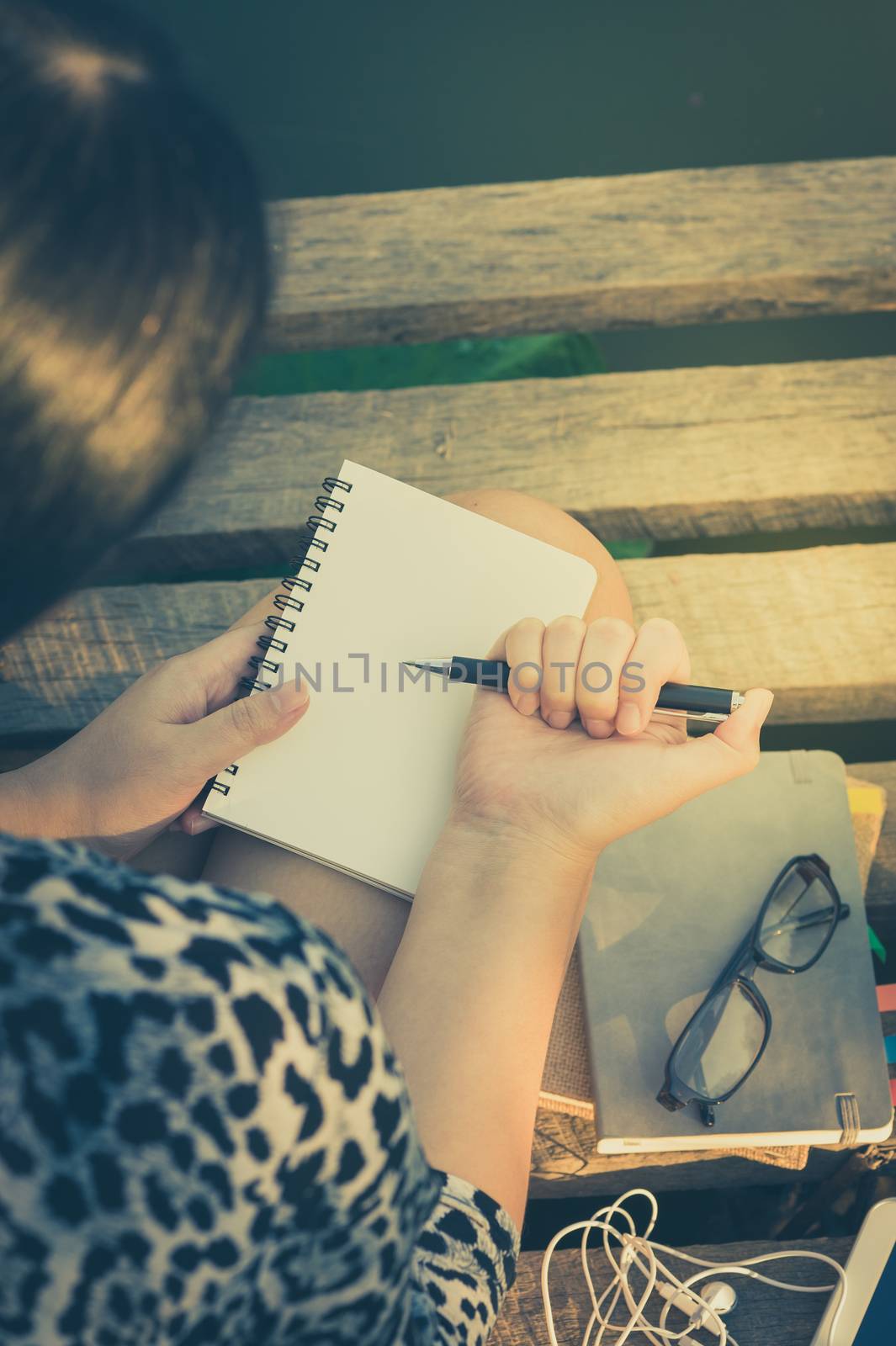 Young hipster woman writing journal on small notebook while sitting on wood bridge in morning time on weekend with vintage filter effect