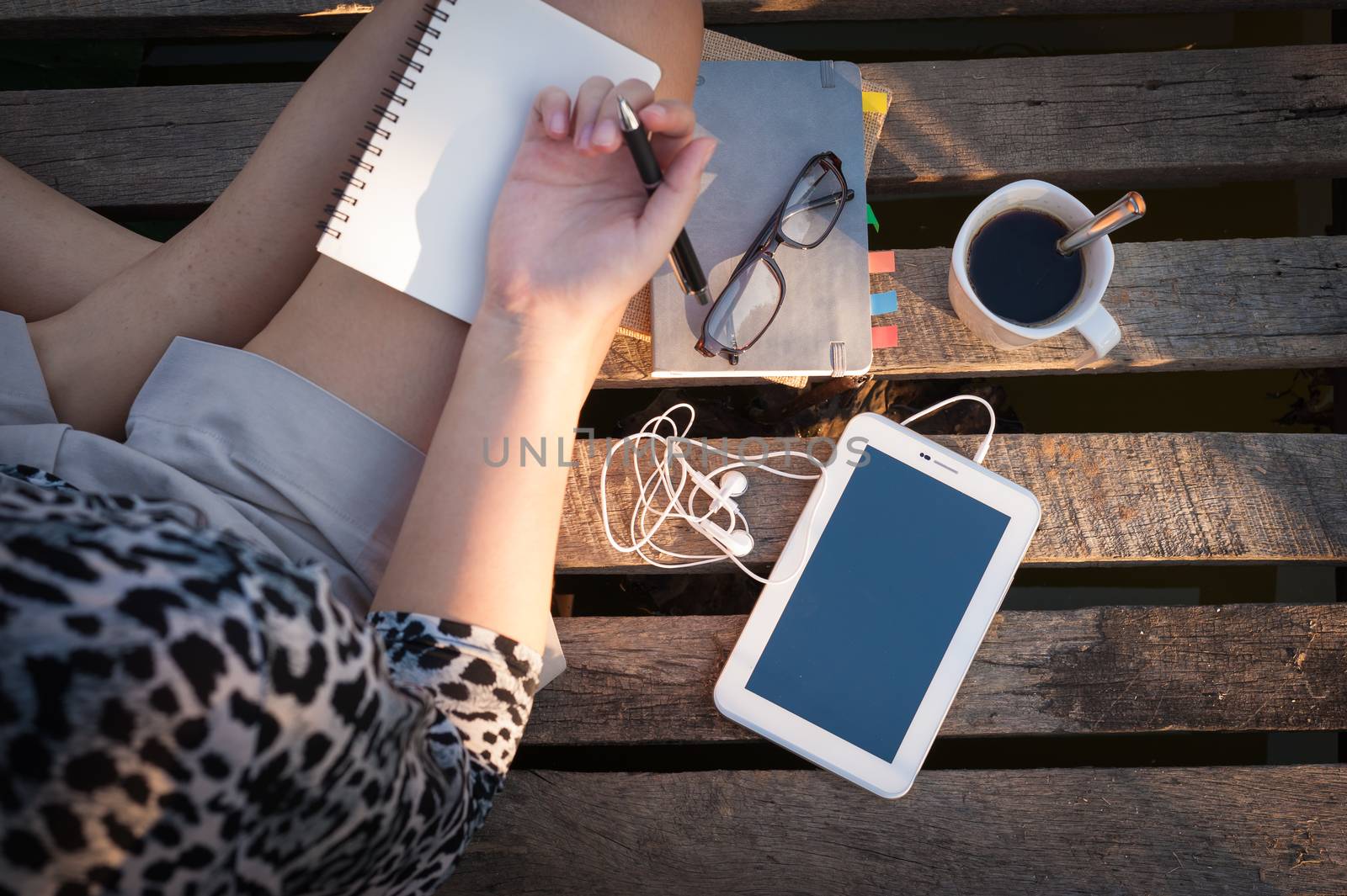 Small tablet pc is put down on wood bridge beside young woman, coffee cup, glasses, and notebooks in early morning time, focus on small tablet pc and notebooks