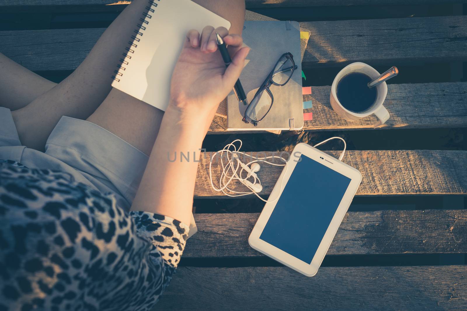 Small tablet pc is put down on wood bridge beside young woman, coffee cup, glasses, and notebooks in early morning time, focus on small tablet pc and notebooks with vintage filer effect