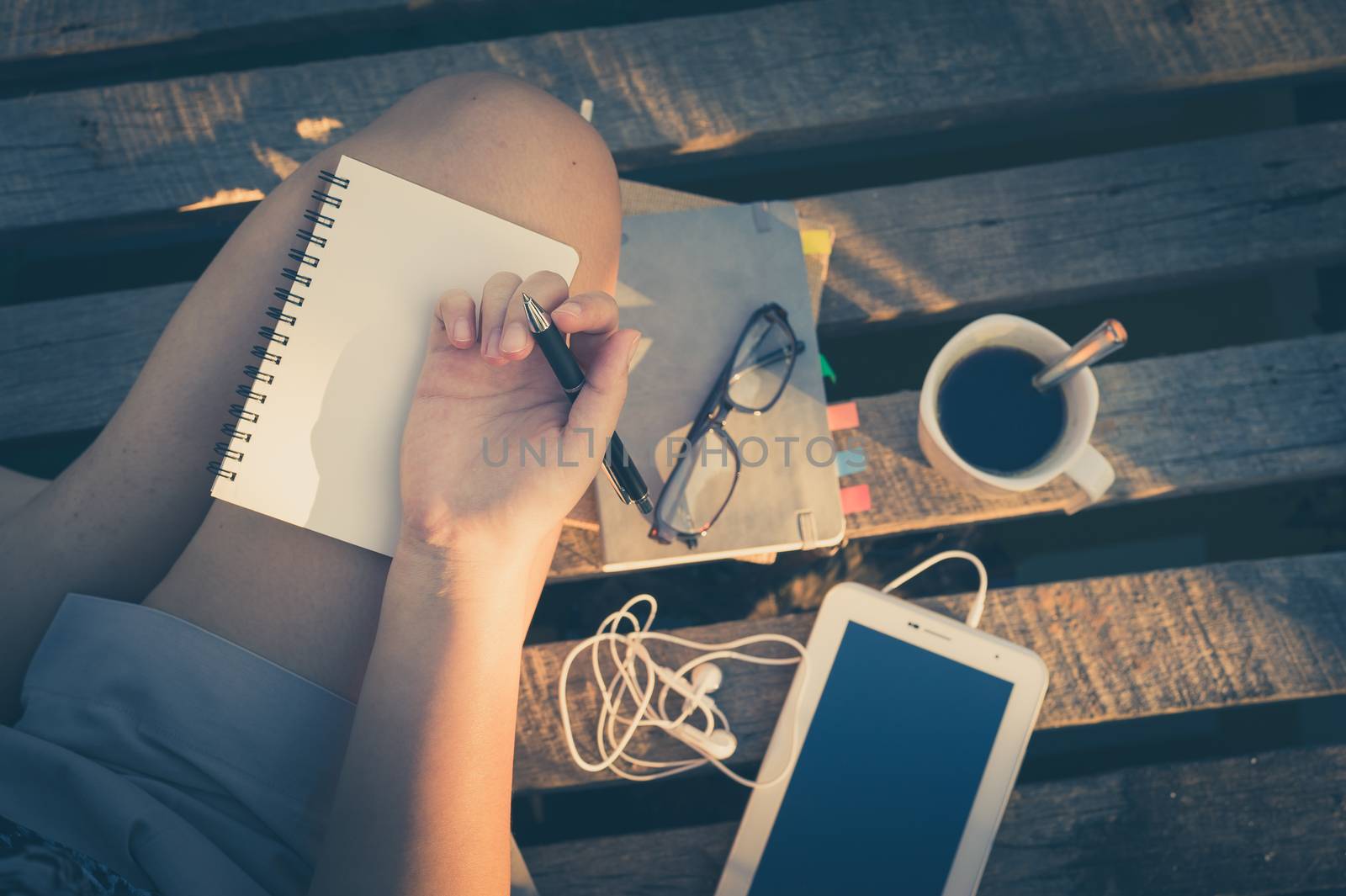 Young hipster woman writing journal on small notebook while sitting on wood litter in early morning time on weekend with vintage filter effect
