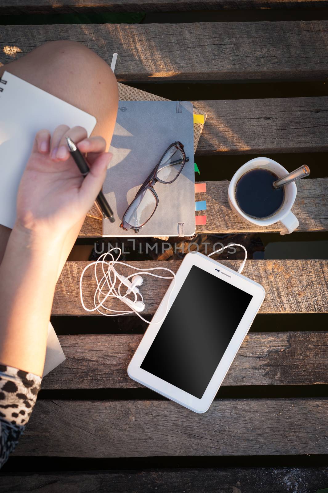 Small tablet pc is put down on wood bridge beside young woman, coffee cup, glasses, and notebooks on morning time