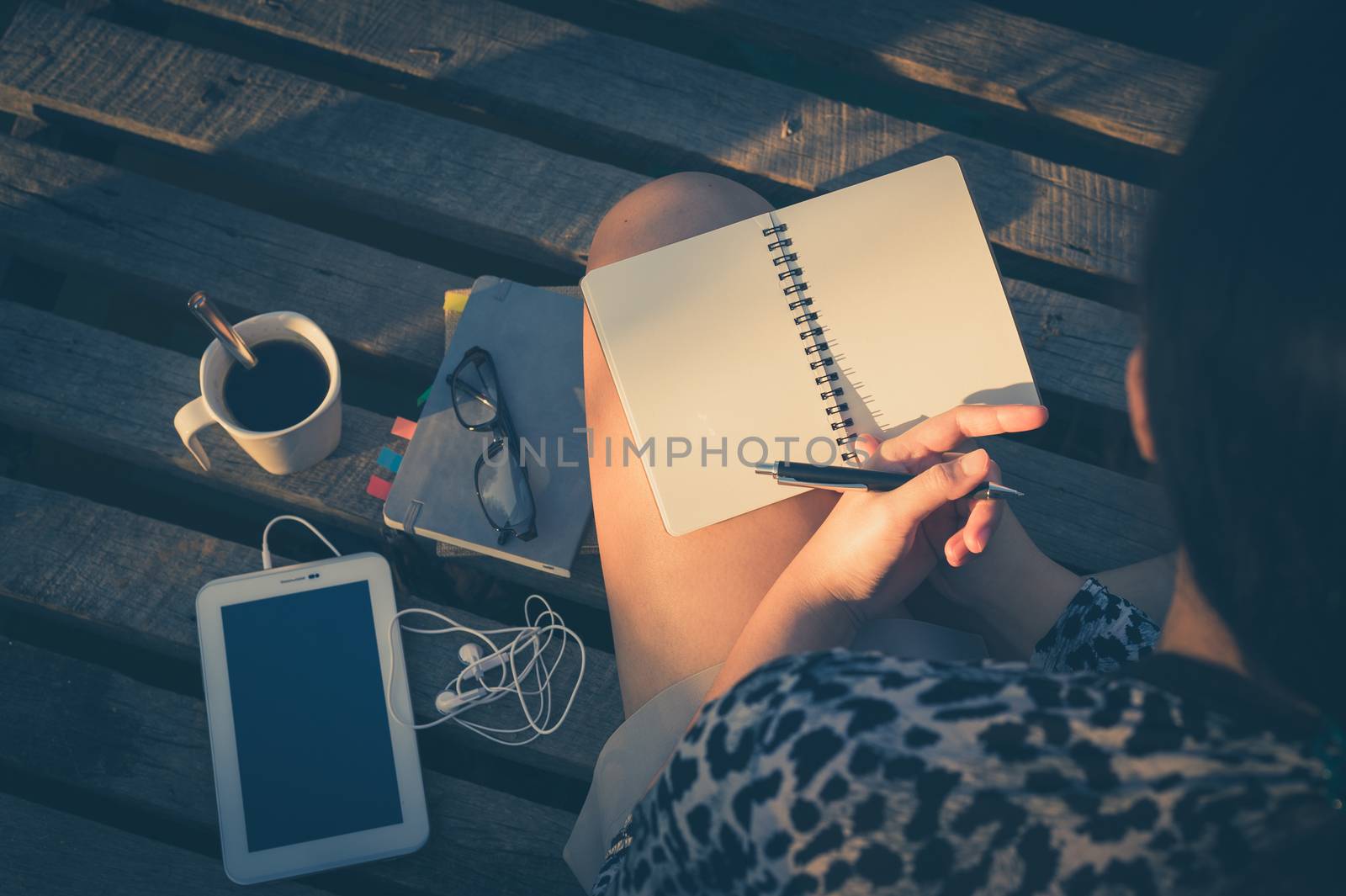 Young hipster woman writing journal on small notebook while sitting on wood bridge in early morning time on weekend with high contrast sun lighting and vintage filter effect