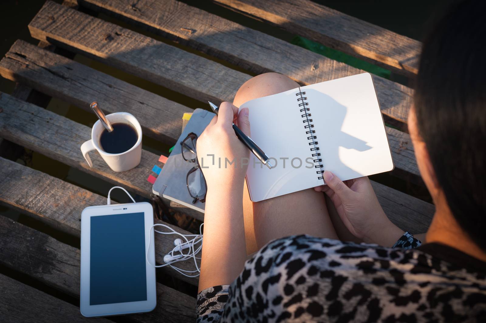 Young hipster woman writing journal on small notebook while sitting on wood bridge in early morning time on weekend with high contrast sun lighting