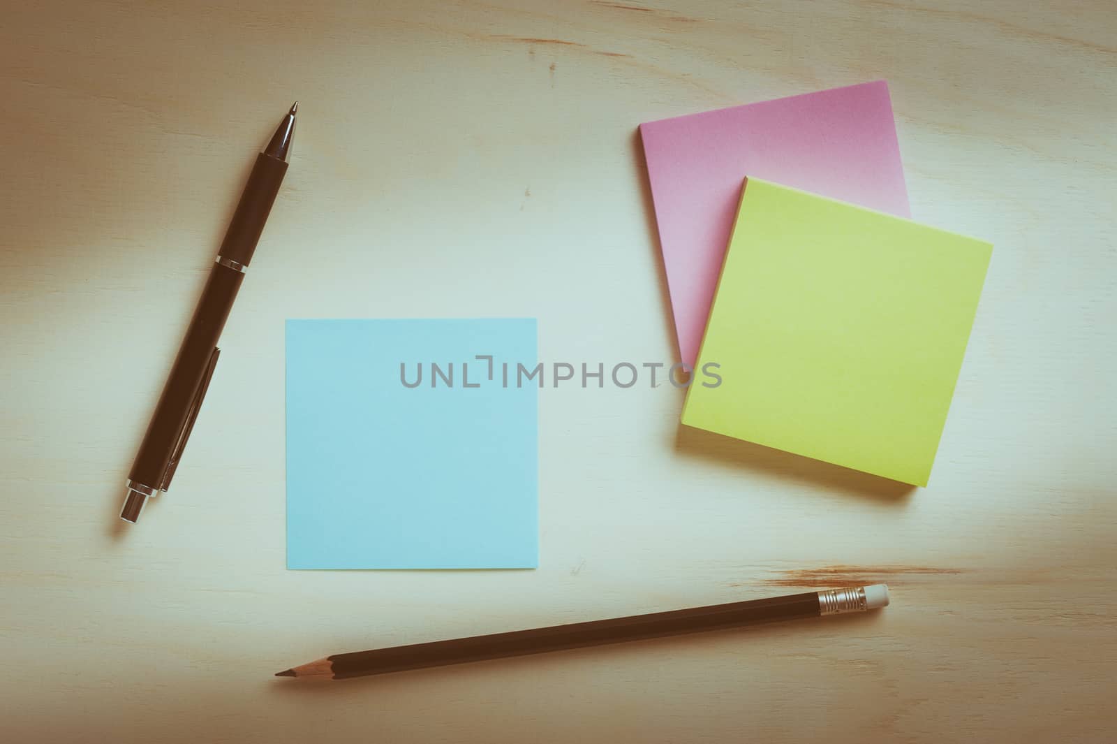 Sticky paper including blue, green, and pink color with blank area for text or message stick on wood table with pen and pencil, processed with vintage filter effect