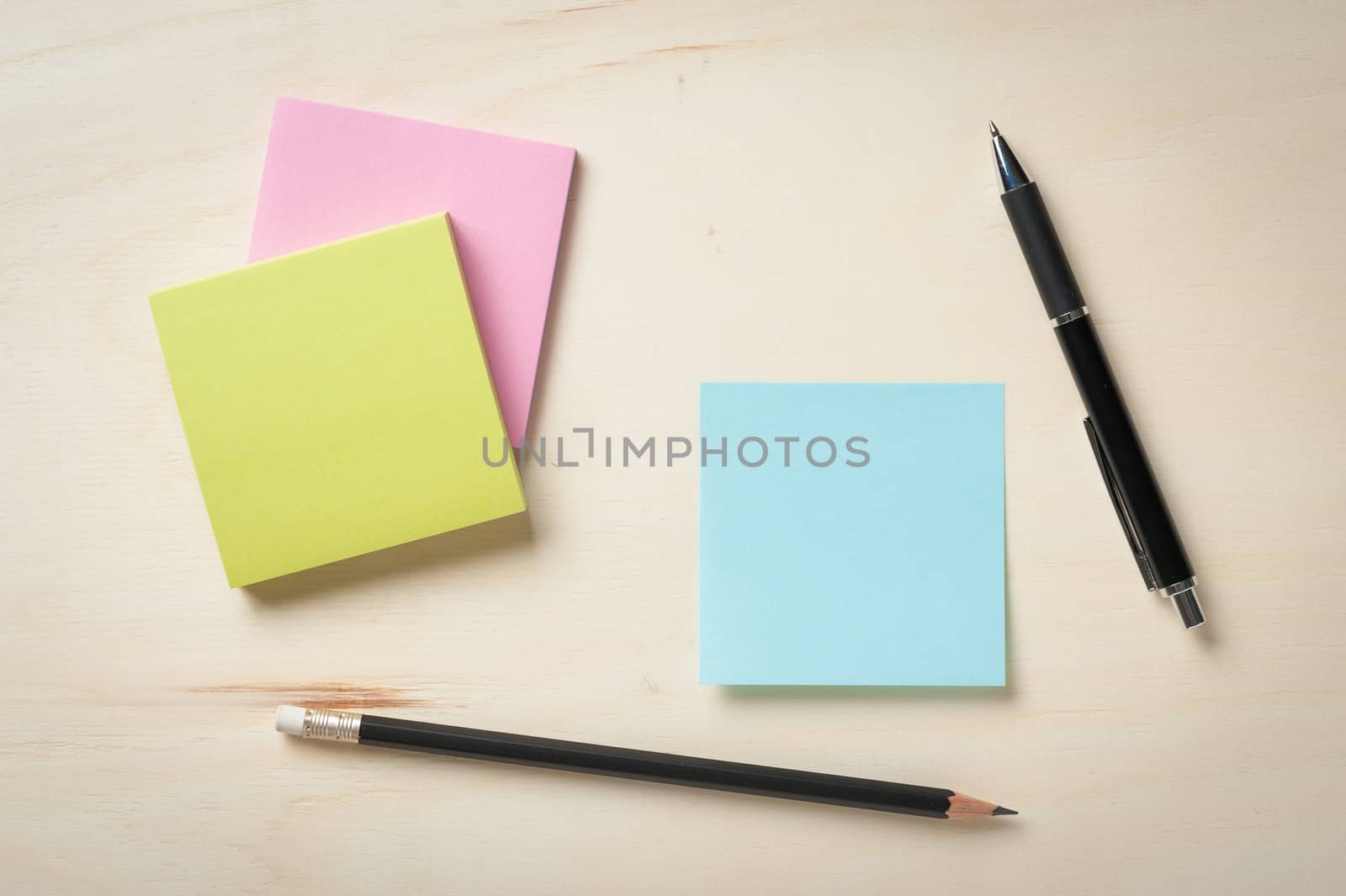 Sticky paper including blue, green, and pink color with blank area for text or message stick on wood table with pen and pencil
