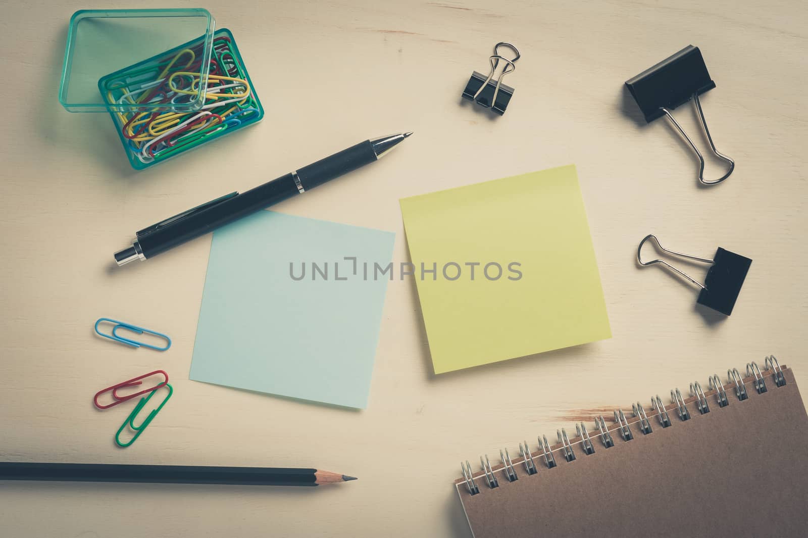 Sticky paper including blue, and green color with blank area for text or message stick on wood table with pen, pencil, paper clips, and notebook in morning time with vintage filter effect