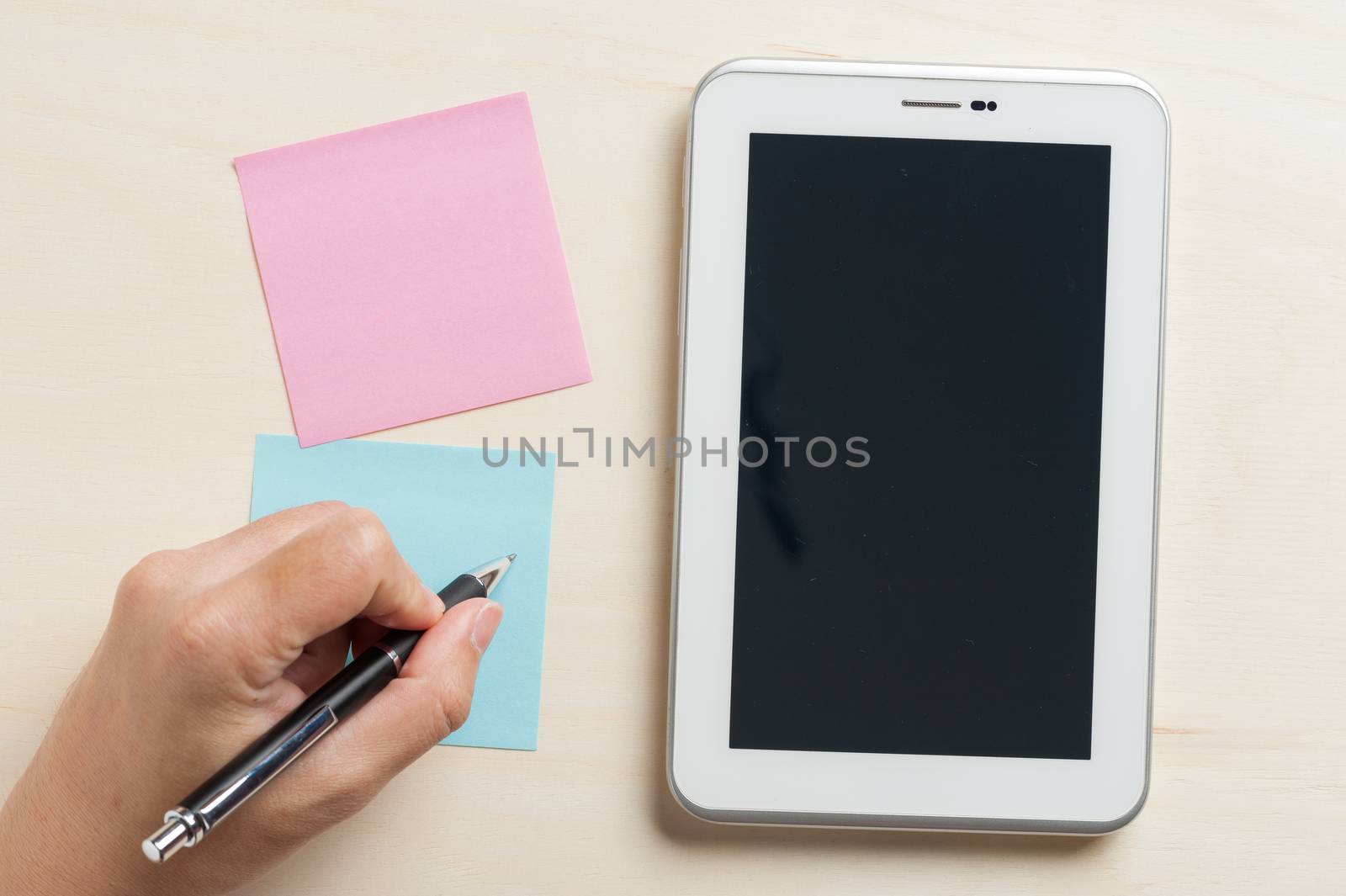 Young man writing with left hand on blue sticky paper beside small tablet pc with blank area on touchscreen on wood table