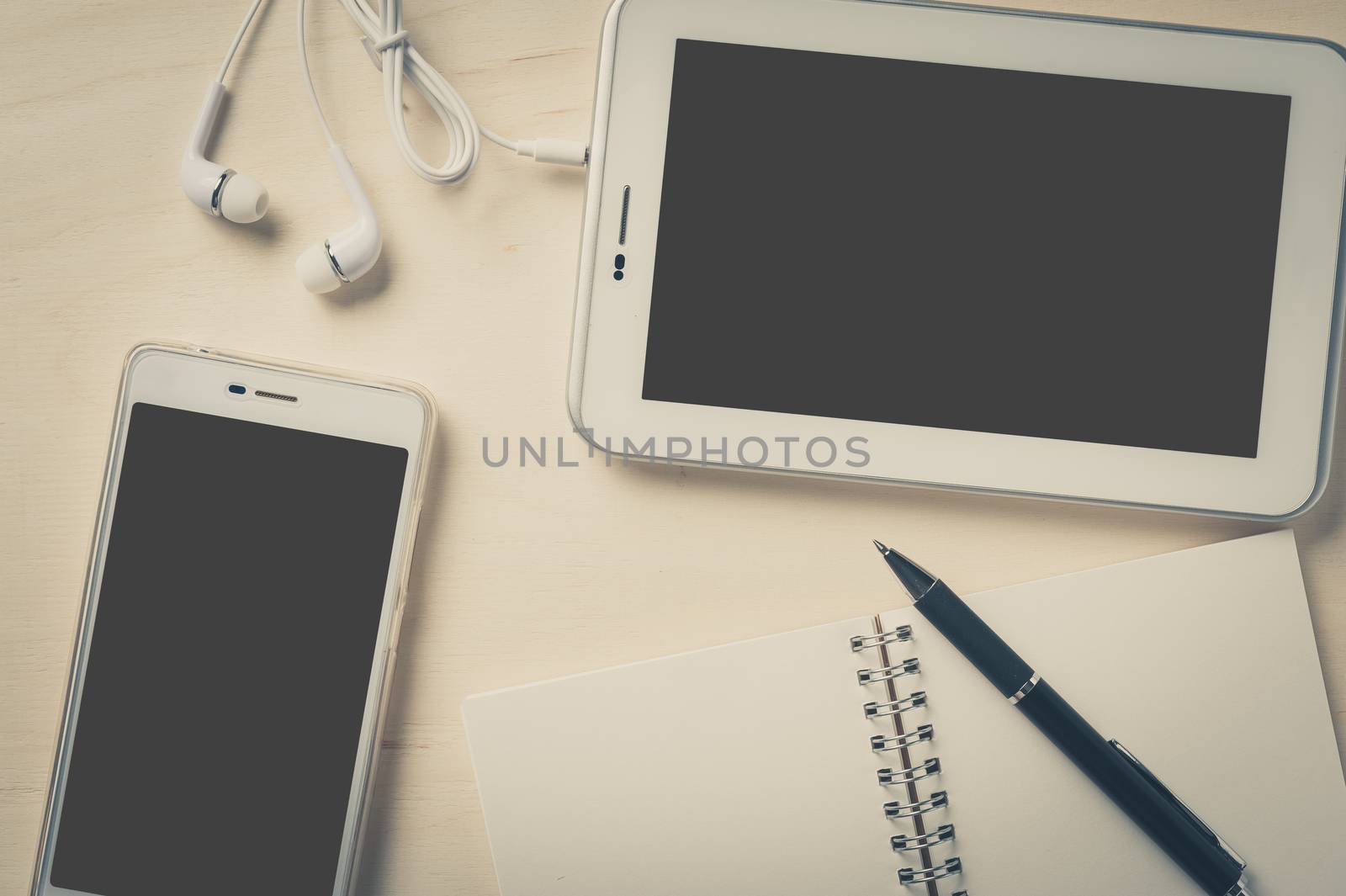 Small tablet pc with blank area on touchscreen, smart phone, in ear headset, notebook, and pen on wood table with vintage filter effect