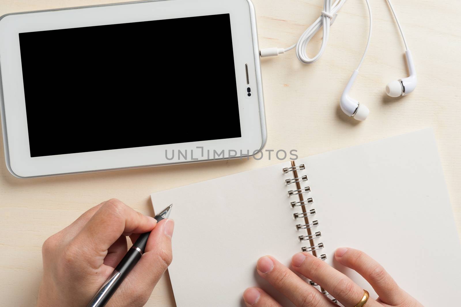 Young man hand writing on notebook beside small tablet pc and in ear headset on wood table