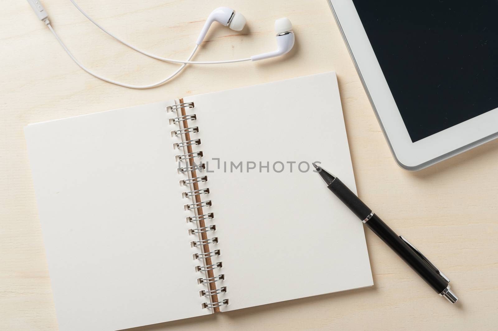 Notebook with blank area for text or message and pen beside small tablet pc and in ear headset on wood table