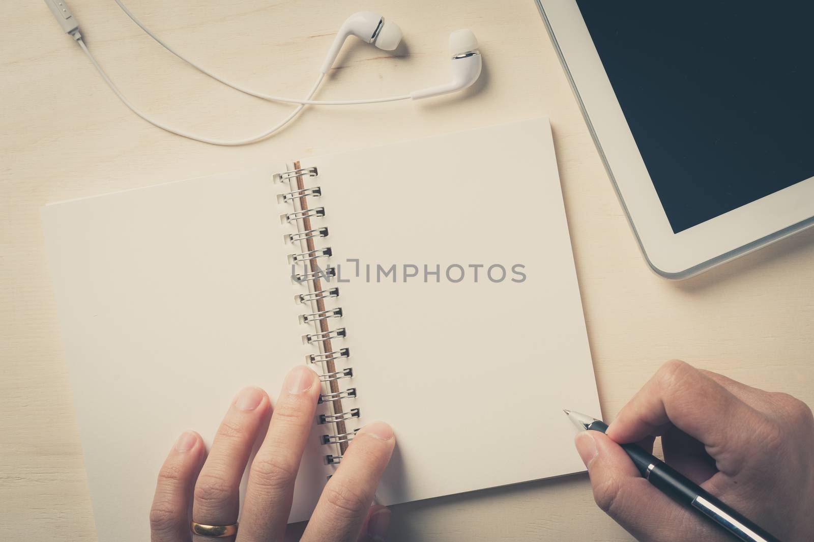 Young man right hand writing on notebook beside small tablet pc and in ear headset on wood table with vintage filter effect