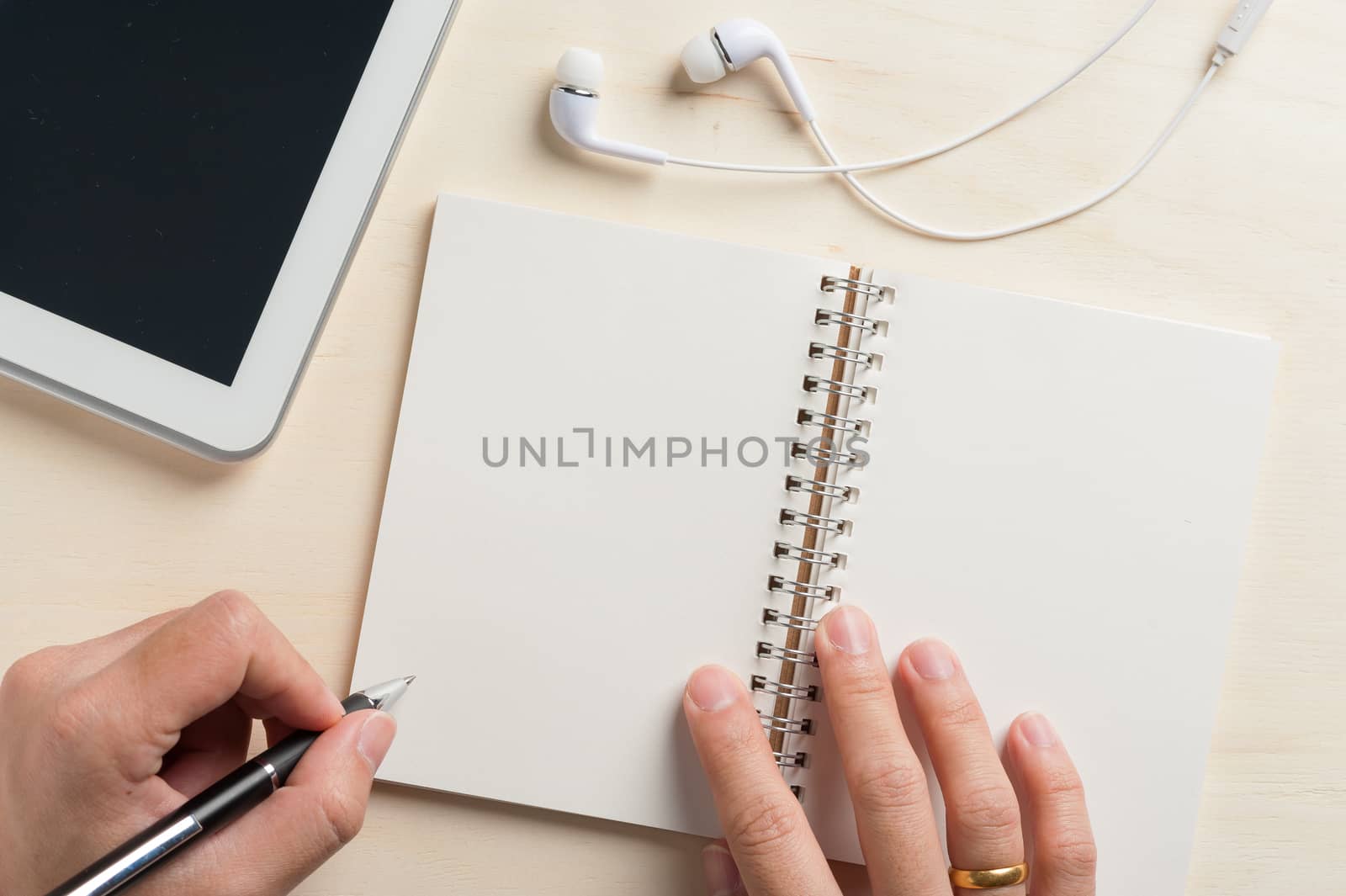Young man left hand writing on notebook beside small tablet pc and in ear headset on wood table