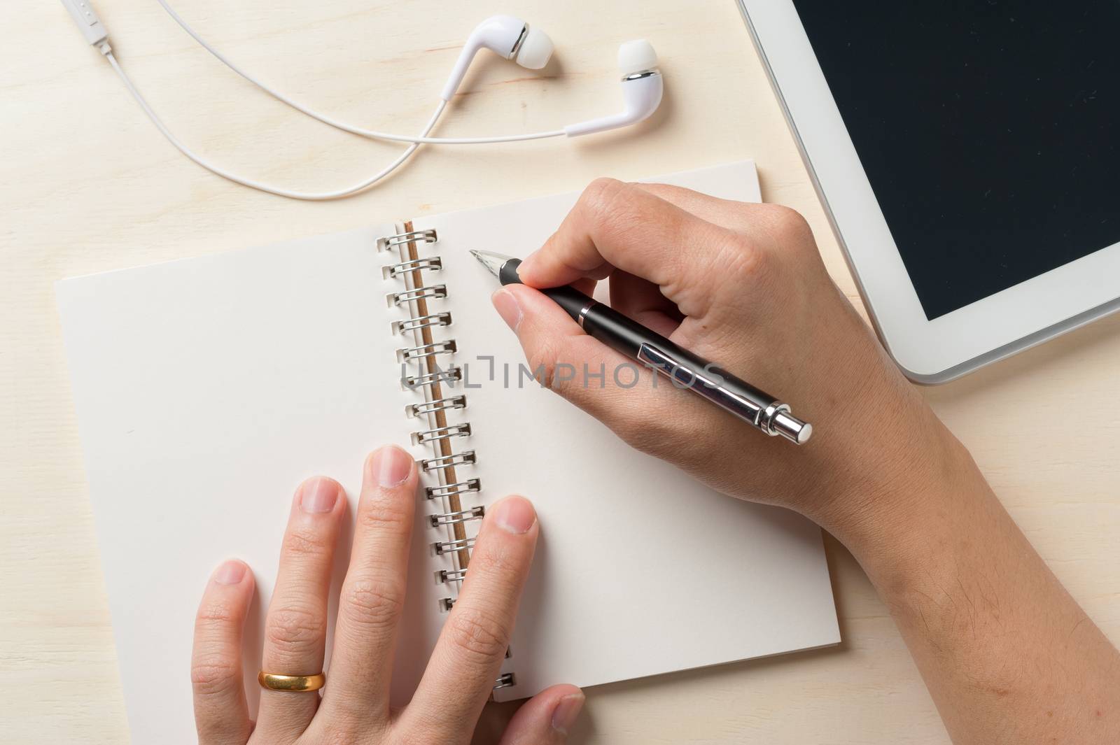 Young man right hand writing on notebook beside small tablet pc and in ear headset on wood table