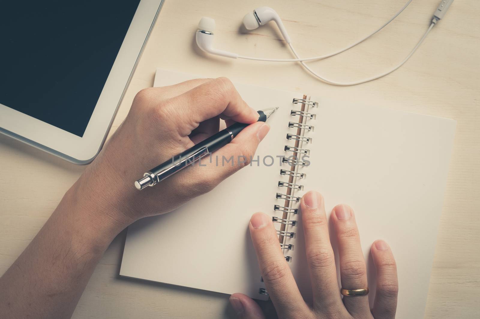 Young man left hand writing on notebook beside small tablet pc and in ear headset on wood table with vintage filter effect