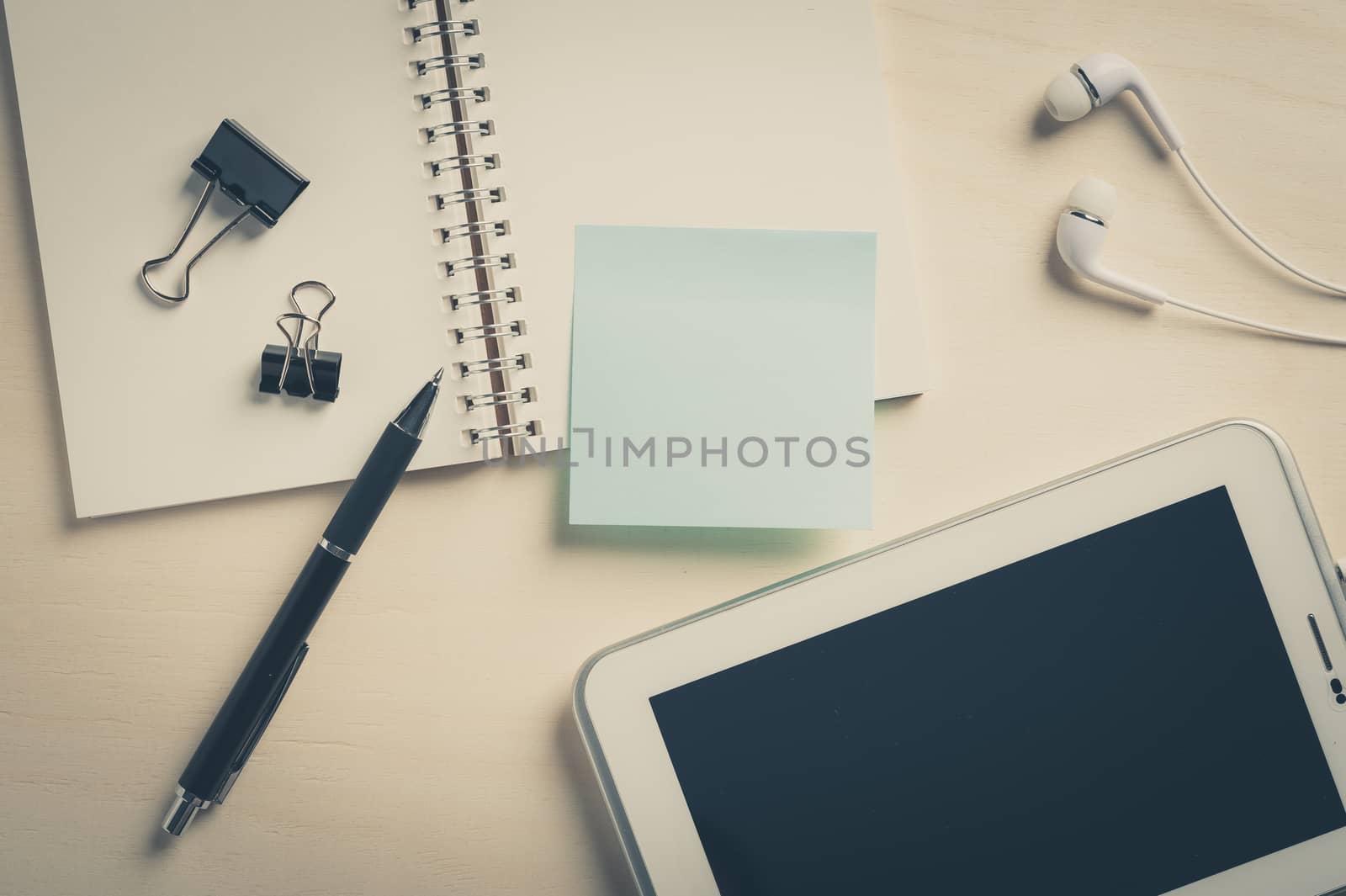 Sticky paper with blank area for text or message, opened notebook,and pen beside small tablet pc and in ear headset on wood table with vintage filter effect