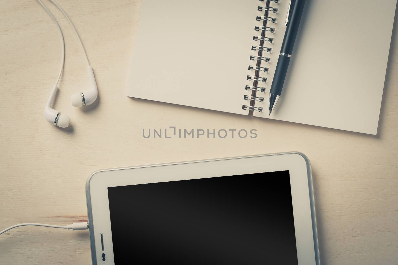 Notebook, pen, small tablet pc, and in ear headset on wood table with vintage filter effect