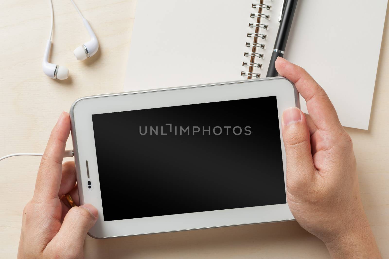 Young man hands holding small tablet pc screen on wood table with in ear headset and notebook beside