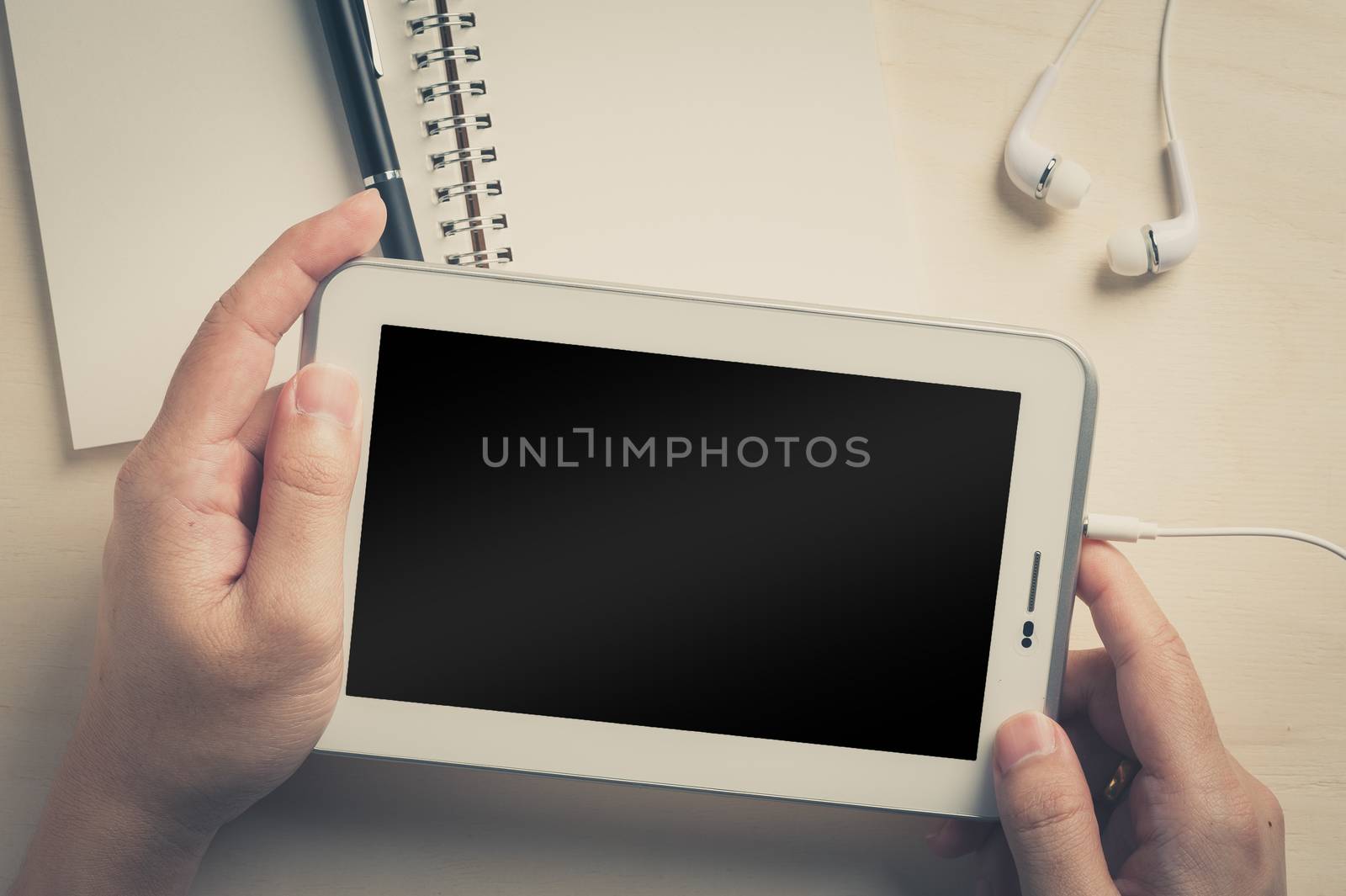 Young man hands holding small tablet pc screen on wood table with in ear headset and notebook beside with vintage filter effect