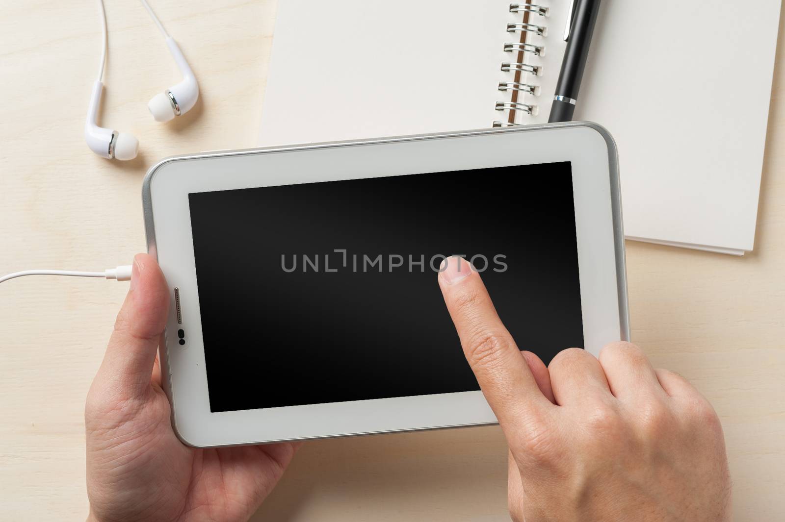 Young man right hand touching on small tablet pc screen on wood table with in ear headset and notebook beside