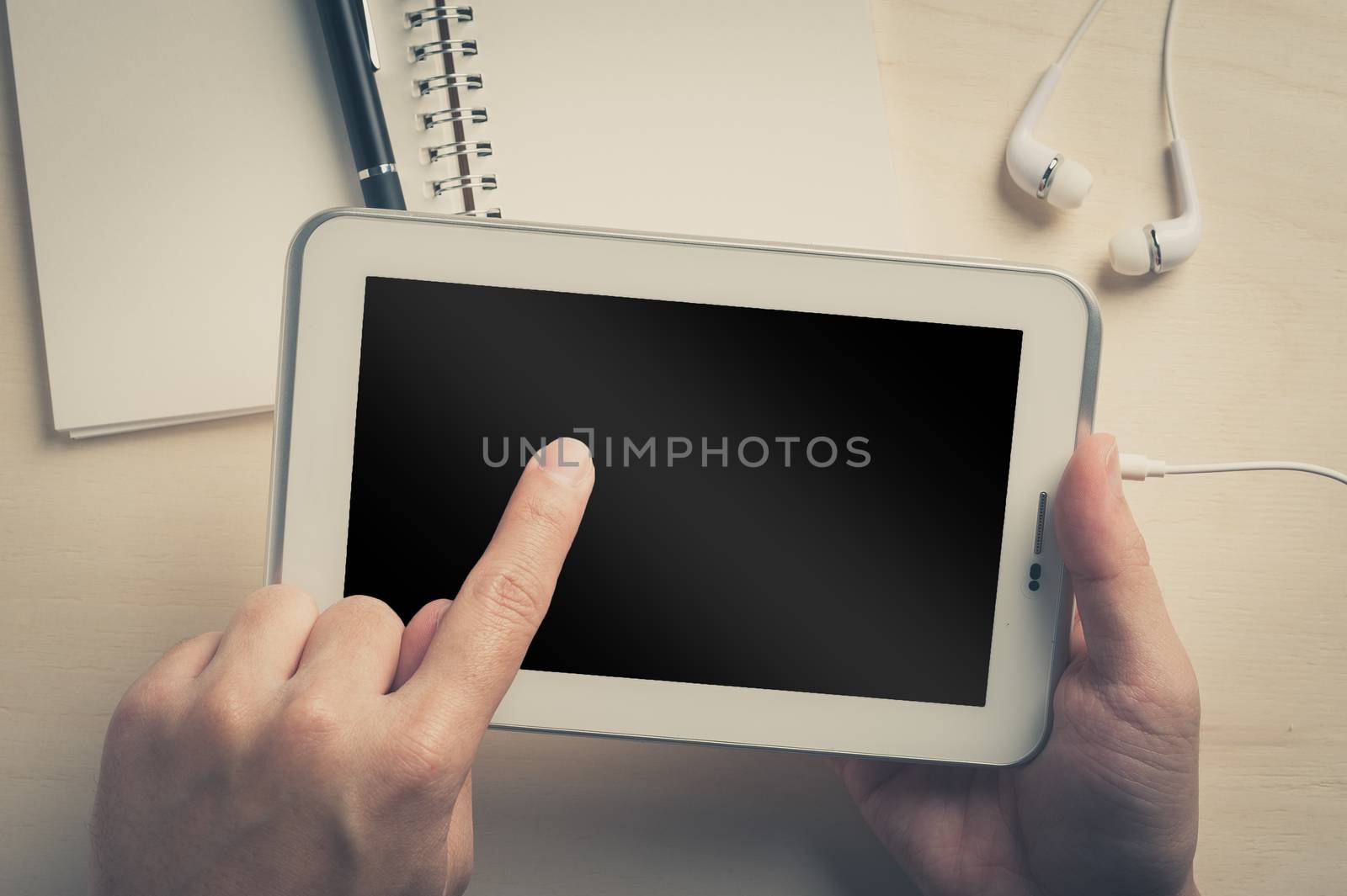 Young man left hand touching on small tablet pc screen on wood table with in ear headset and notebook beside with vintage filter effect