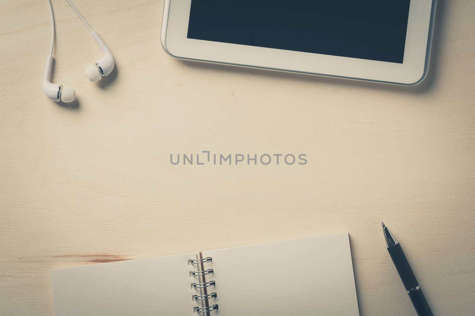 Notebook, pen, small tablet pc, and in ear headset on wood table with vintage filter effect